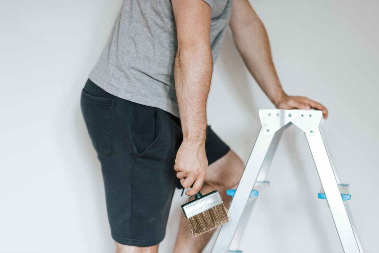 hero-img Vue de côté d’un décorateur masculin anonyme en tenue décontractée préparant la peinture des murs d’une pièce claire, debout sur un escabeau